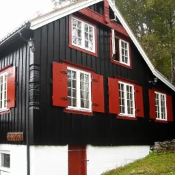 Czarny drewniany dom z czerwonymi okiennicami i białymi ramami okiennymi, widok z zewnątrz, częściowo widoczne schody wejściowe.