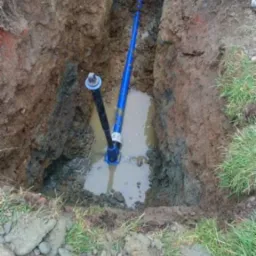 Wykopany rów wypełniony wodą z widocznymi niebieskimi rurami i czarną rurą pionową z zaworem, sugerujący etap prac ziemnych związanych z instalacją podziemną.