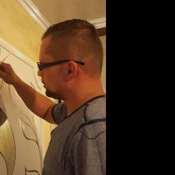 Decco - Mężczyzna w okularach maluje ozdobny wzór złotą farbą na ścianie w ciepłym oświetleniu.