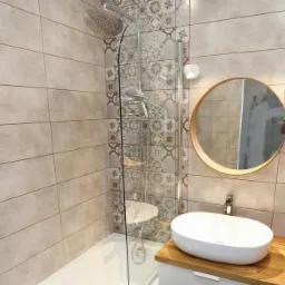 Nowoczesna łazienka z beżowymi płytkami imitującymi drewno, kabiną prysznicową ze szklaną ścianką zdobioną wzorzystymi kaflami, okrągłym lustrem w drewnianej ramie nad białą umywalką nablatową...