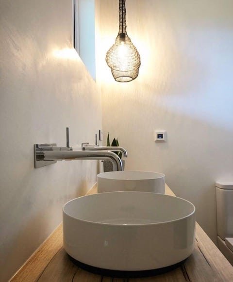 Nowoczesna łazienka z dwoma okrągłymi umywalkami nablatowymi na drewnianym blacie, chromowaną armaturą ścienną i dekoracyjną lampą wiszącą z metalowej siatki.