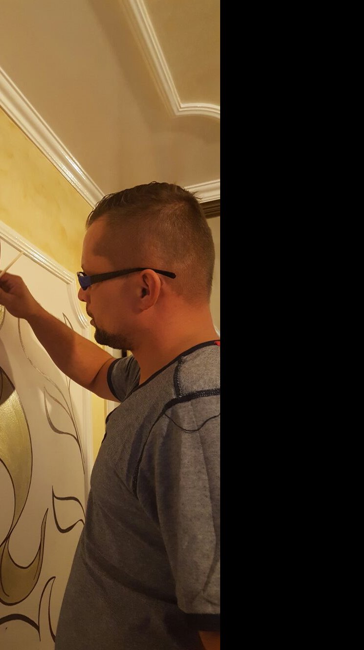 Mężczyzna w okularach maluje ozdobny wzór złotą farbą na ścianie w ciepłym oświetleniu.