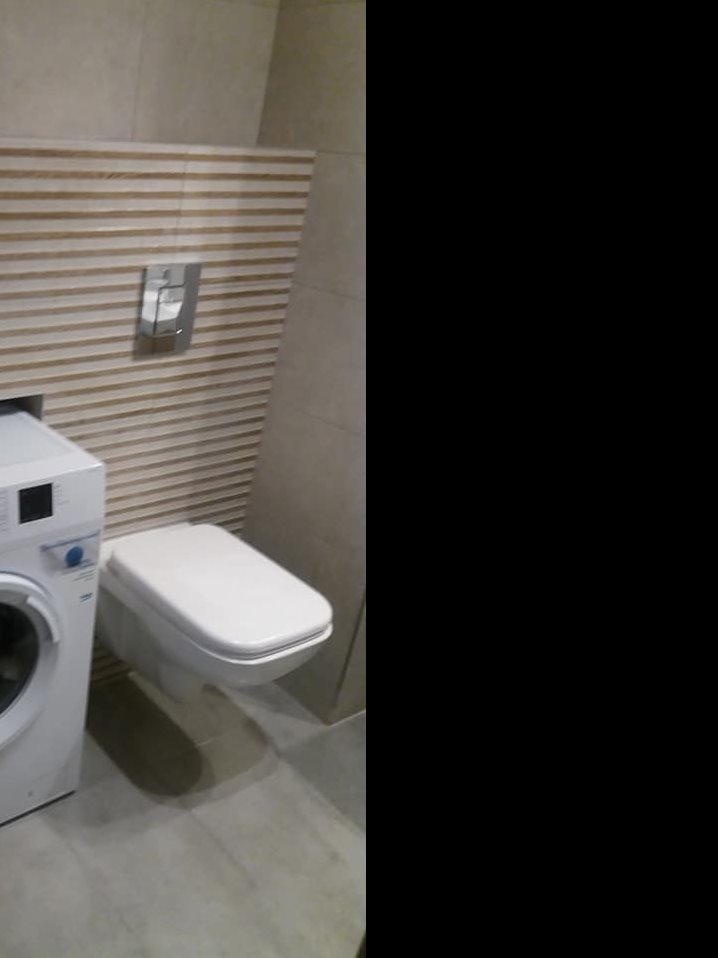 Nowoczesna toaleta podwieszana z białą deską, obok pralka, na ścianie płytki imitujące drewno w odcieniach beżu i szarości, widoczny chromowany przycisk spłuczki.