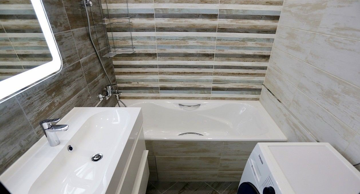 Nowoczesna łazienka z umywalką na szafce, wanną z chromowanymi uchwytami i pralką, wykończona płytkami imitującymi drewno w różnych odcieniach szarości, brązu i błękitu.