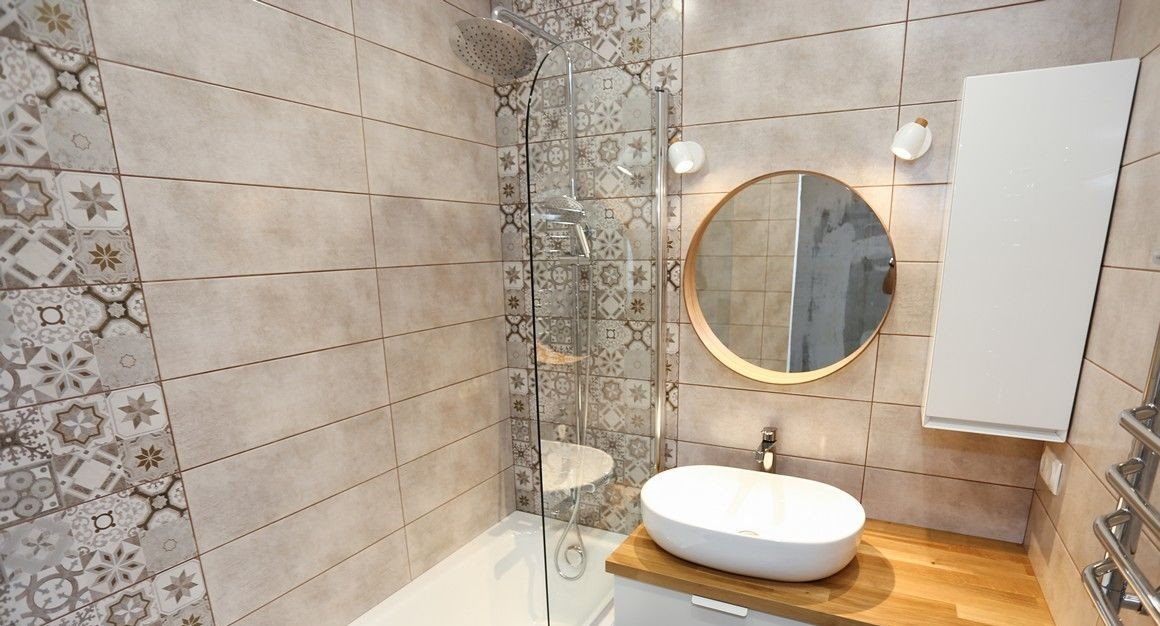 Nowoczesna łazienka z beżowymi płytkami imitującymi drewno, kabiną prysznicową ze szklaną ścianką zdobioną wzorzystymi kaflami, okrągłym lustrem w drewnianej ramie nad białą umywalką nablatową...