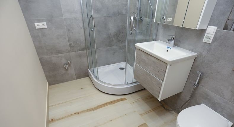 Nowoczesna łazienka z szarymi płytkami ściennymi, kabiną prysznicową narożną, umywalką z szafką w kolorze drewna i jasną podłogą imitującą drewno.