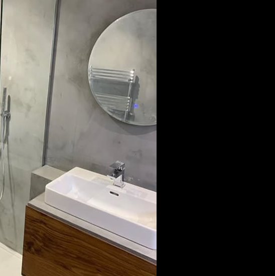 Nowoczesna łazienka z umywalką nablatową na szafce z drewna, okrągłe lustro z podświetleniem i grzejnik drabinkowy na ścianie wykończonej szarym betonem architektonicznym.