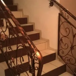 Wewnętrzna balustrada schodowa z metalu, zdobiona motywami roślinnymi, w kolorze ciemnej miedzi, zamontowana przy schodach z białymi stopniami i brązowymi podstopnicami.