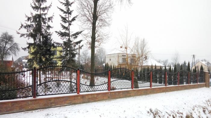 Czarne, metalowe ogrodzenie z czerwonymi słupkami na niskiej, ceglanej podmurówce, otoczone śniegiem w pochmurny zimowy dzień. W tle widoczne domy i drzewa.