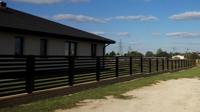 Nowoczesne ogrodzenie posesji z czarnych, poziomych paneli na ceglanej podmurówce, widok z zewnątrz na tle domu i słupów wysokiego napięcia.