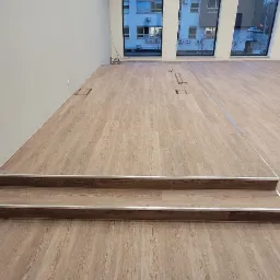 Jasna podłoga z paneli drewnopodobnych w dużym pomieszczeniu biurowym, widoczne dwa stopnie schodów z metalowymi listwami, okna z widokiem na budynek mieszkalny.