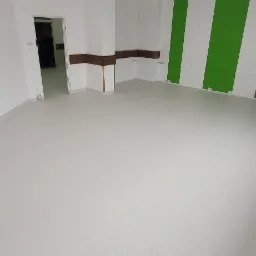 Jasna, pusta sala z nową, jednolitą wykładziną PCV w odcieniach szarości, kontrastująca z zielono-białymi pasami na ścianie i widocznymi lampami sufitowymi.