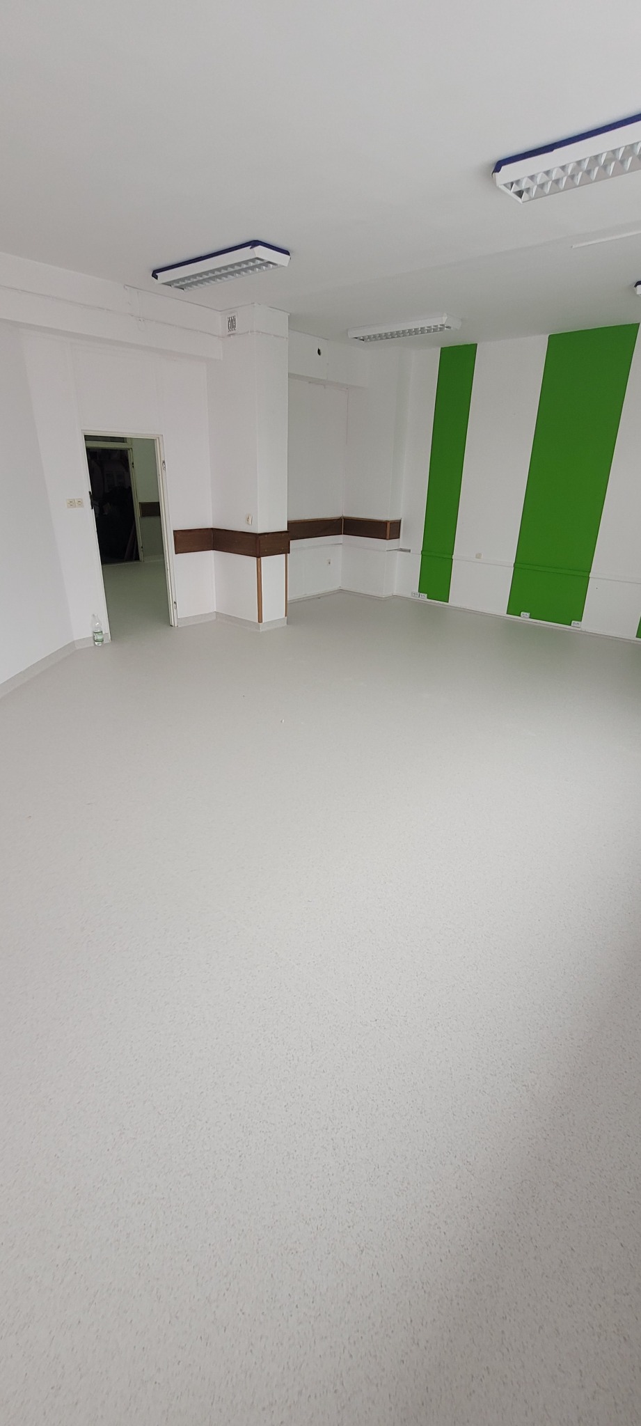 Jasna, pusta sala z nową, jednolitą wykładziną PCV w odcieniach szarości, kontrastująca z zielono-białymi pasami na ścianie i widocznymi lampami sufitowymi.