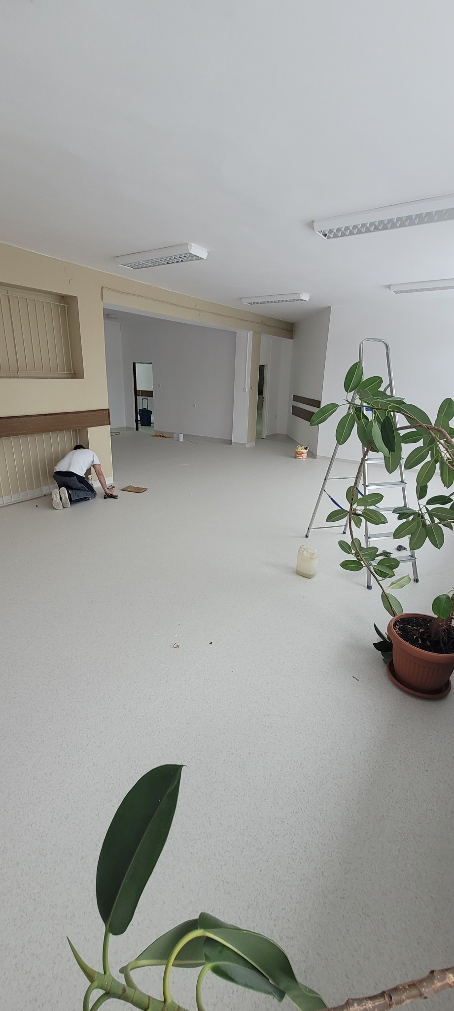 Pracownik na kolanach docina jasną wykładzinę w dużym, pustym pomieszczeniu z widocznymi lampami sufitowymi i roślinami doniczkowymi w rogu.