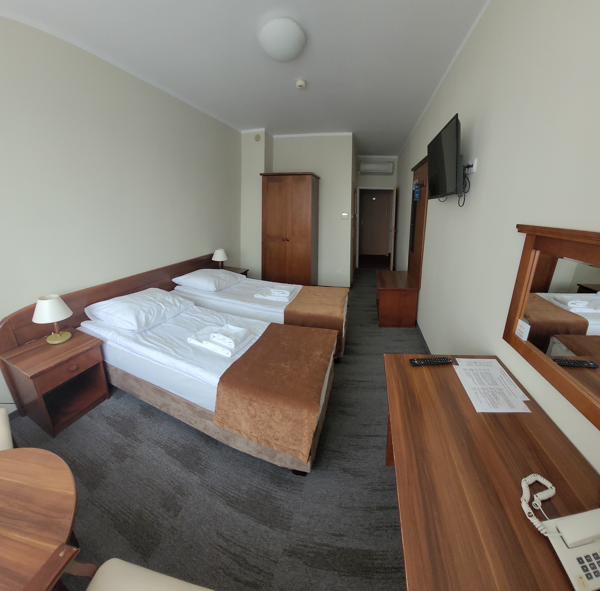 Pokój hotelowy z dwoma łóżkami, szarą wykładziną dywanową, drewnianymi meblami i telewizorem na ścianie.