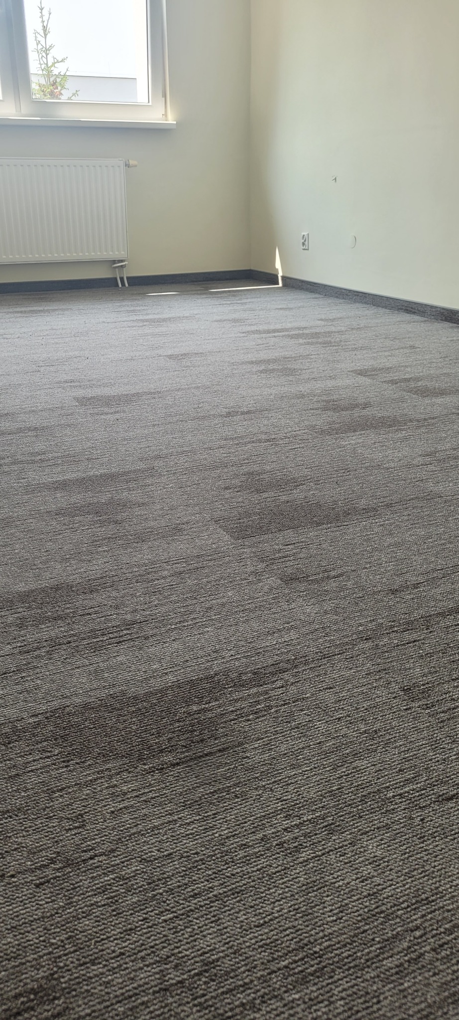 Szara wykładzina dywanowa ułożona w kwadraty z delikatnym wzorem, widok na grzejnik i okno w jasnym pomieszczeniu.