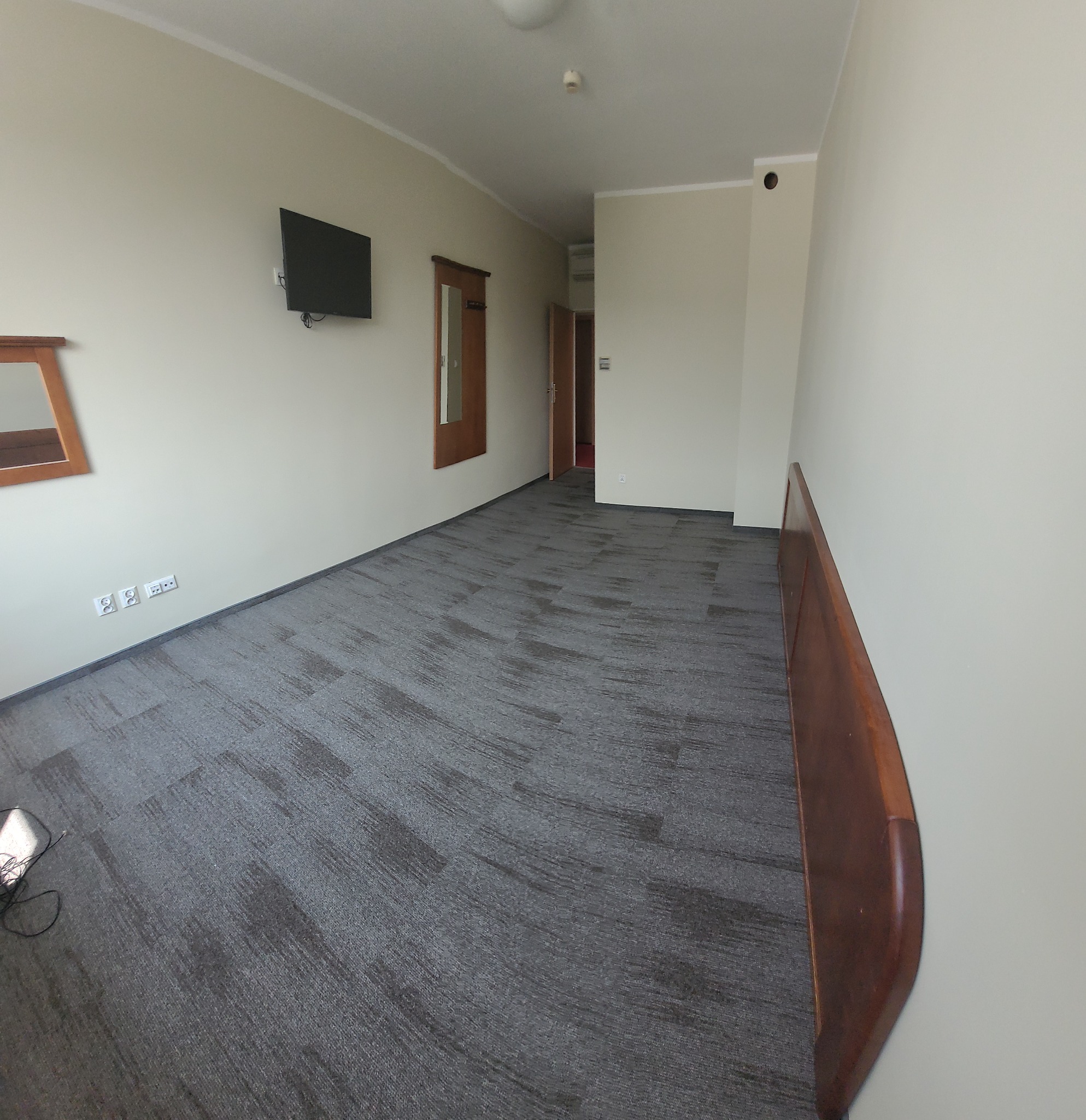 Szara wykładzina dywanowa w pokoju hotelowym, widok na zamontowany telewizor i lustro w drewnianej ramie.