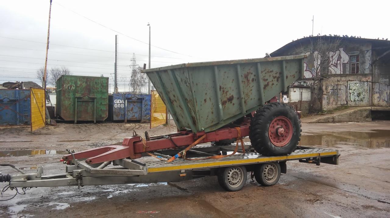 Laweta z przyczepioną skorodowaną przyczepą rolniczą z jednym kołem, zabezpieczona pasami transportowymi, na tle przemysłowego otoczenia z kontenerami i budynkiem pokrytym graffiti.