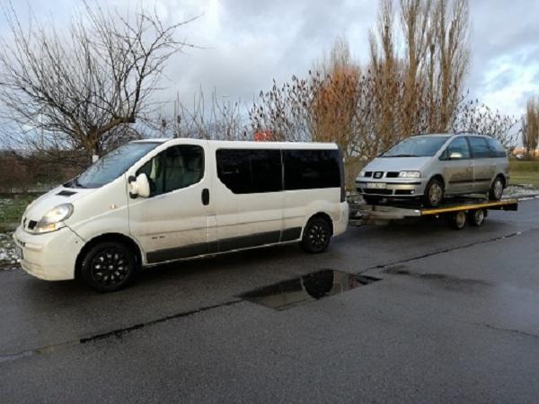 Biały bus z czarnymi felgami ciągnie przyczepę z srebrnym samochodem osobowym na tle drzew bez liści i pochmurnego nieba.