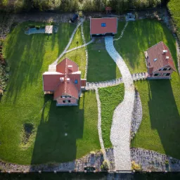 Widok z lotu ptaka na zadbany ogród z trzema budynkami, wybrukowanymi ścieżkami, stawem i równo przystrzyżonym trawnikiem, otoczony ogrodzeniem.