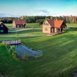 CUDOGRODY.PL Ewa Liberadzka-Cudnoch - Rozległy, zadbany trawnik otacza trzy domy z czerwonymi dachami, mały staw z drewnianym pomostem i saunę w kształcie beczki; widok z lotu ptaka.