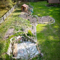 CUDOGRODY.PL Ewa Liberadzka-Cudnoch - Widok z góry na rozległy ogród z domkiem dla dzieci, altaną z grillem, siatką do siatkówki i kamiennym patio z paleniskiem.