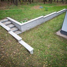 CUDOGRODY.PL Ewa Liberadzka-Cudnoch - Nowoczesny murek oporowy z jasnoszarej kostki przy domu w Świętajnie, zintegrowany ze schodami prowadzącymi na trawnik pokryty opadłymi liśćmi.