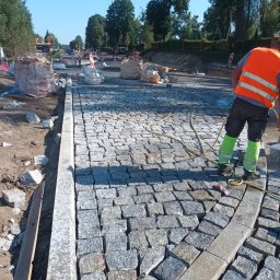 CUDOGRODY.PL Ewa Liberadzka-Cudnoch - Układanie kostki granitowej na drodze. Widoczny fragment ułożonej nawierzchni, pracownik w kamizelce odblaskowej i worki z materiałem budowlanym w tle.