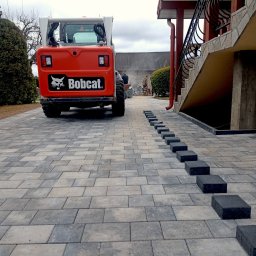 CUDOGRODY.PL Ewa Liberadzka-Cudnoch - Ułożona kostka brukowa w odcieniach szarości z pomarańczowym Bobcatem w tle. Widoczne czarne kostki czekające na ułożenie wzdłuż ściany budynku.