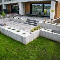 CUDOGRODY.PL Ewa Liberadzka-Cudnoch - Nowoczesny ogród z tarasem wyłożonym szarą kostką, białymi kamieniami w rabatach i świeżo posadzoną trawą. Widok na budynek mieszkalny i ulicę.