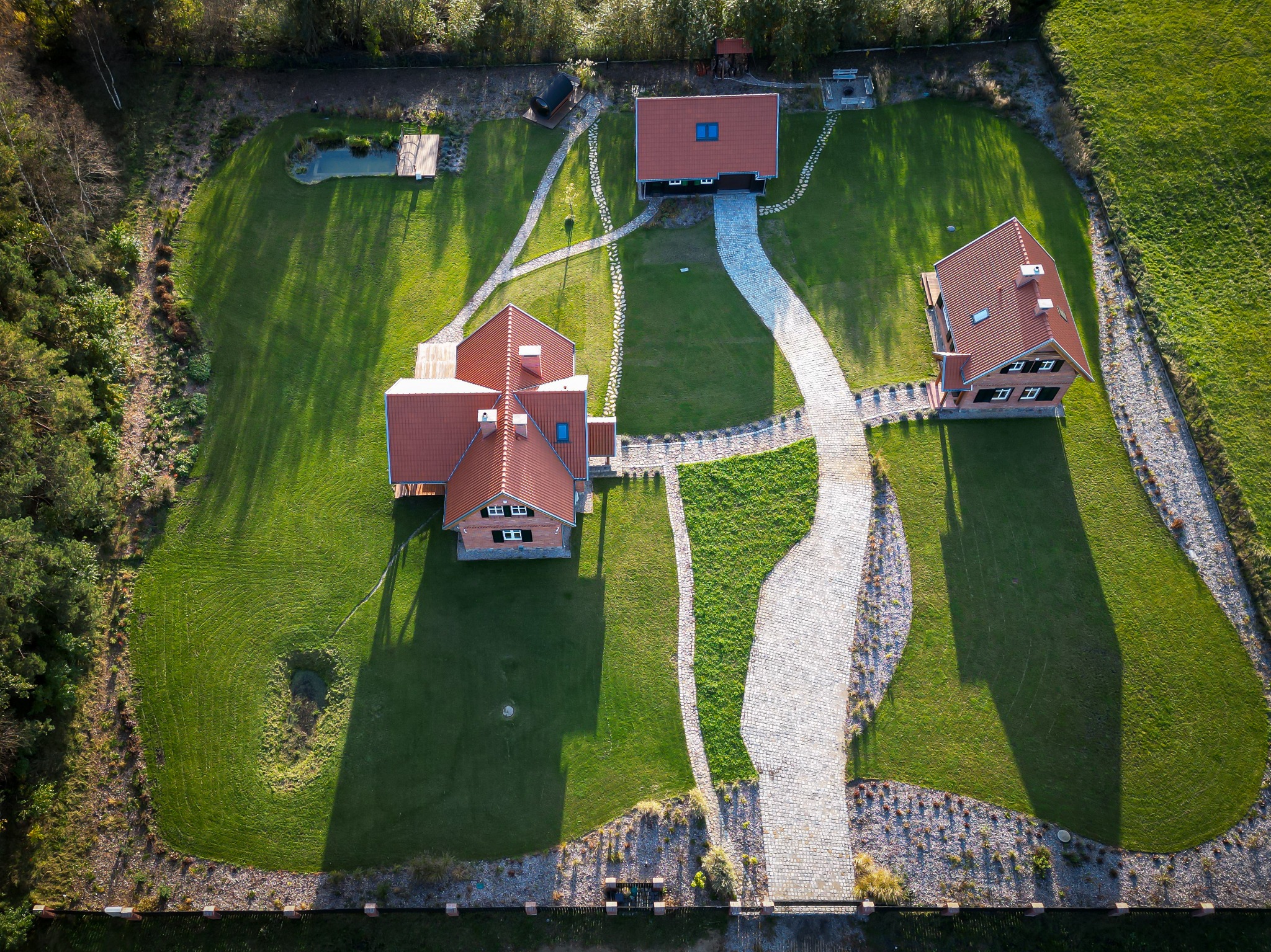 Widok z lotu ptaka na zadbany ogród z trzema budynkami, wybrukowanymi ścieżkami, stawem i równo przystrzyżonym trawnikiem, otoczony ogrodzeniem.