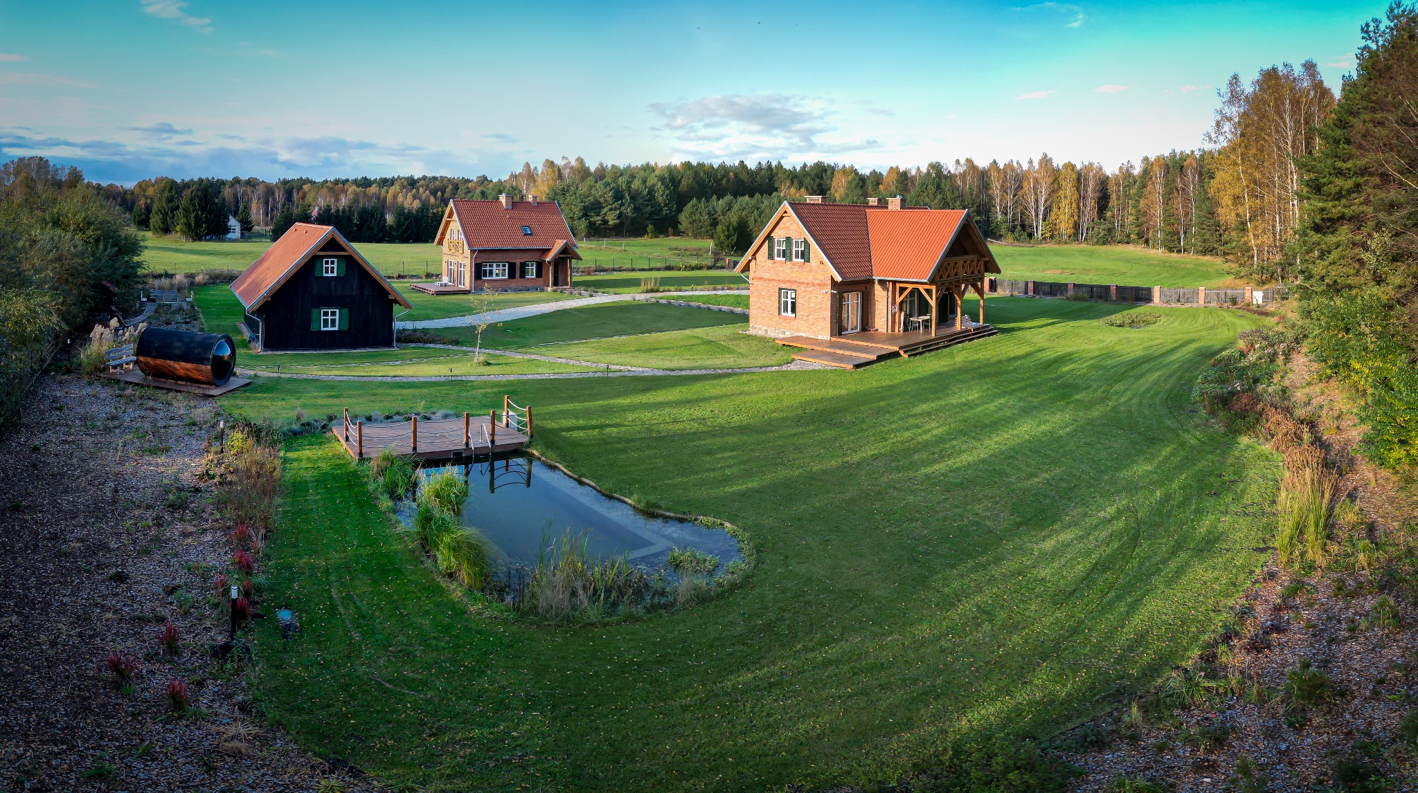 Rozległy, zadbany trawnik otacza trzy domy z czerwonymi dachami, mały staw z drewnianym pomostem i saunę w kształcie beczki; widok z lotu ptaka.