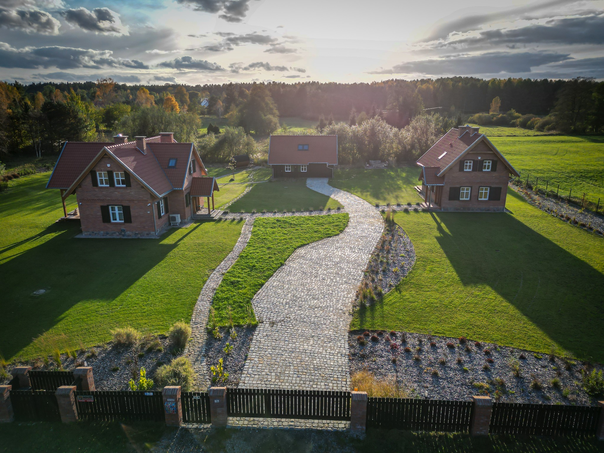 Widok z lotu ptaka na posesję z trzema domami, brukowaną ścieżką z kostki granitowej, zadbanym trawnikiem i ozdobnymi rabatami.