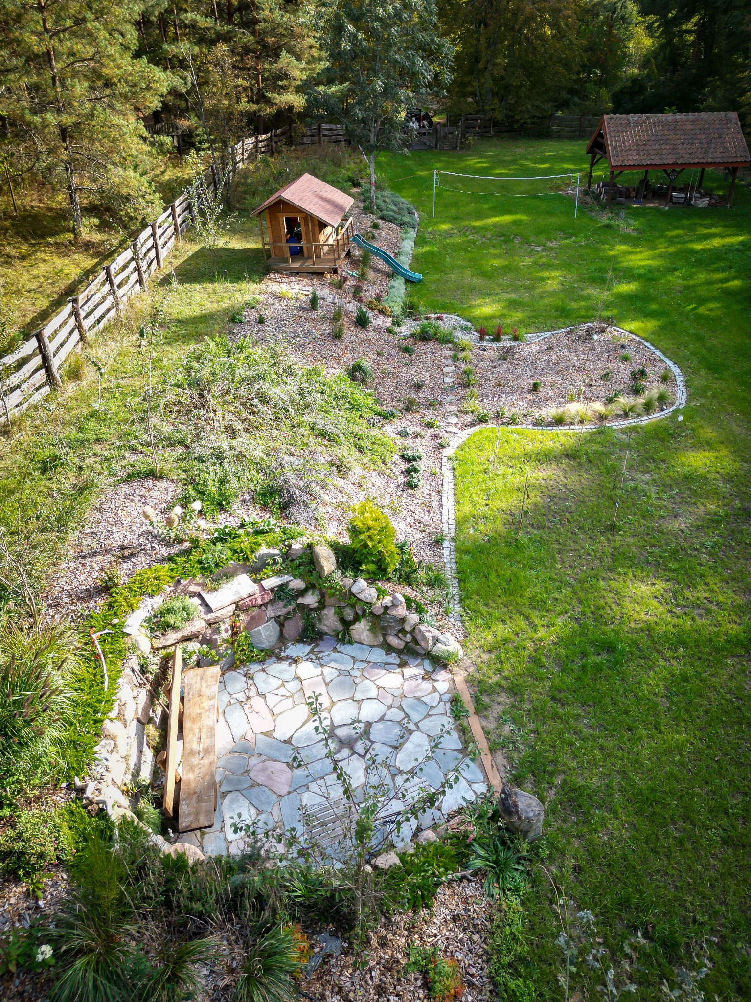 Widok z góry na rozległy ogród z domkiem dla dzieci, altaną z grillem, siatką do siatkówki i kamiennym patio z paleniskiem.