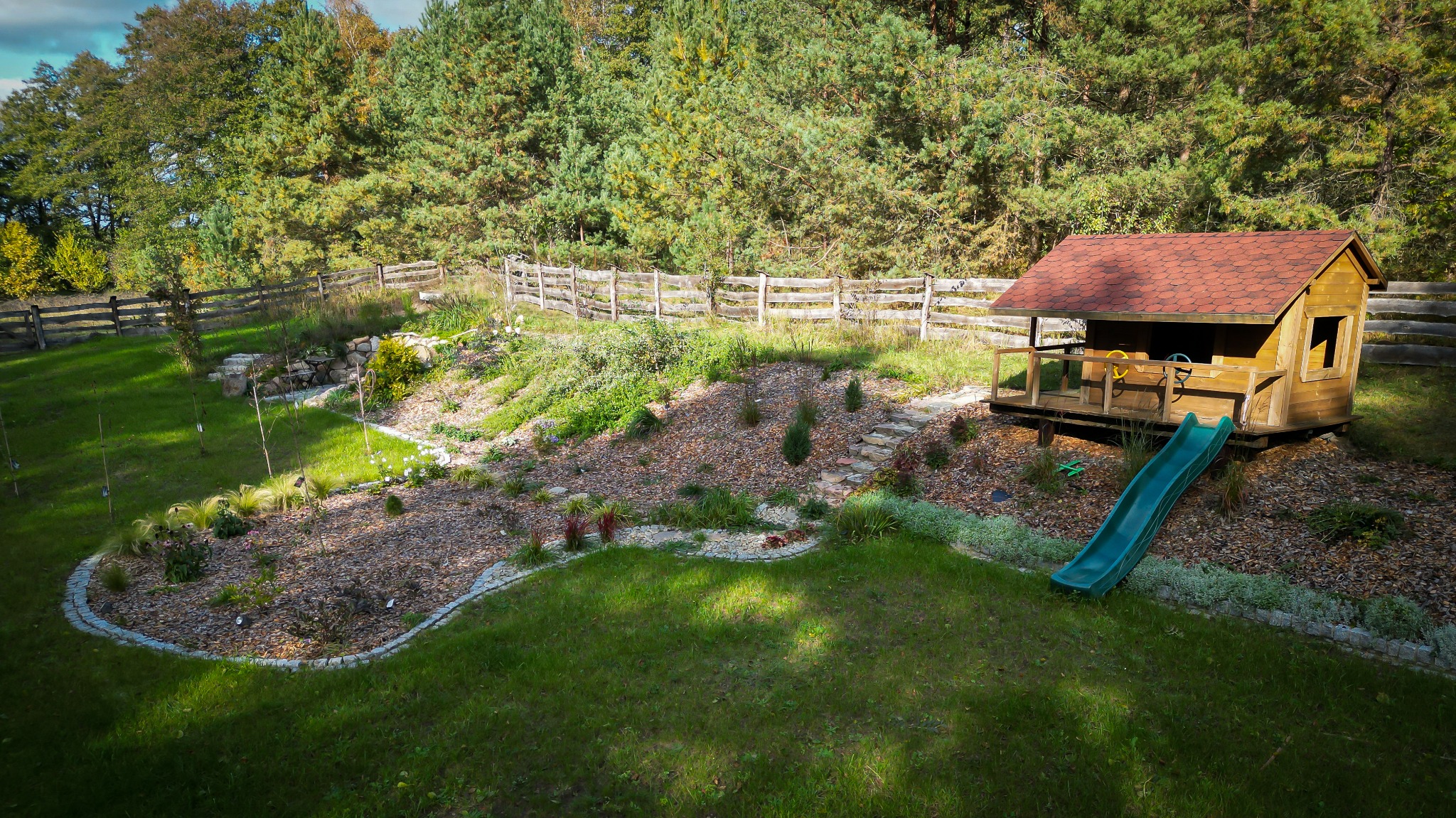 Widok z góry na zagospodarowany ogród z drewnianym domkiem dla dzieci na podwyższeniu, zjeżdżalnią i rabatami wysypanymi korą, otoczony drewnianym płotem.