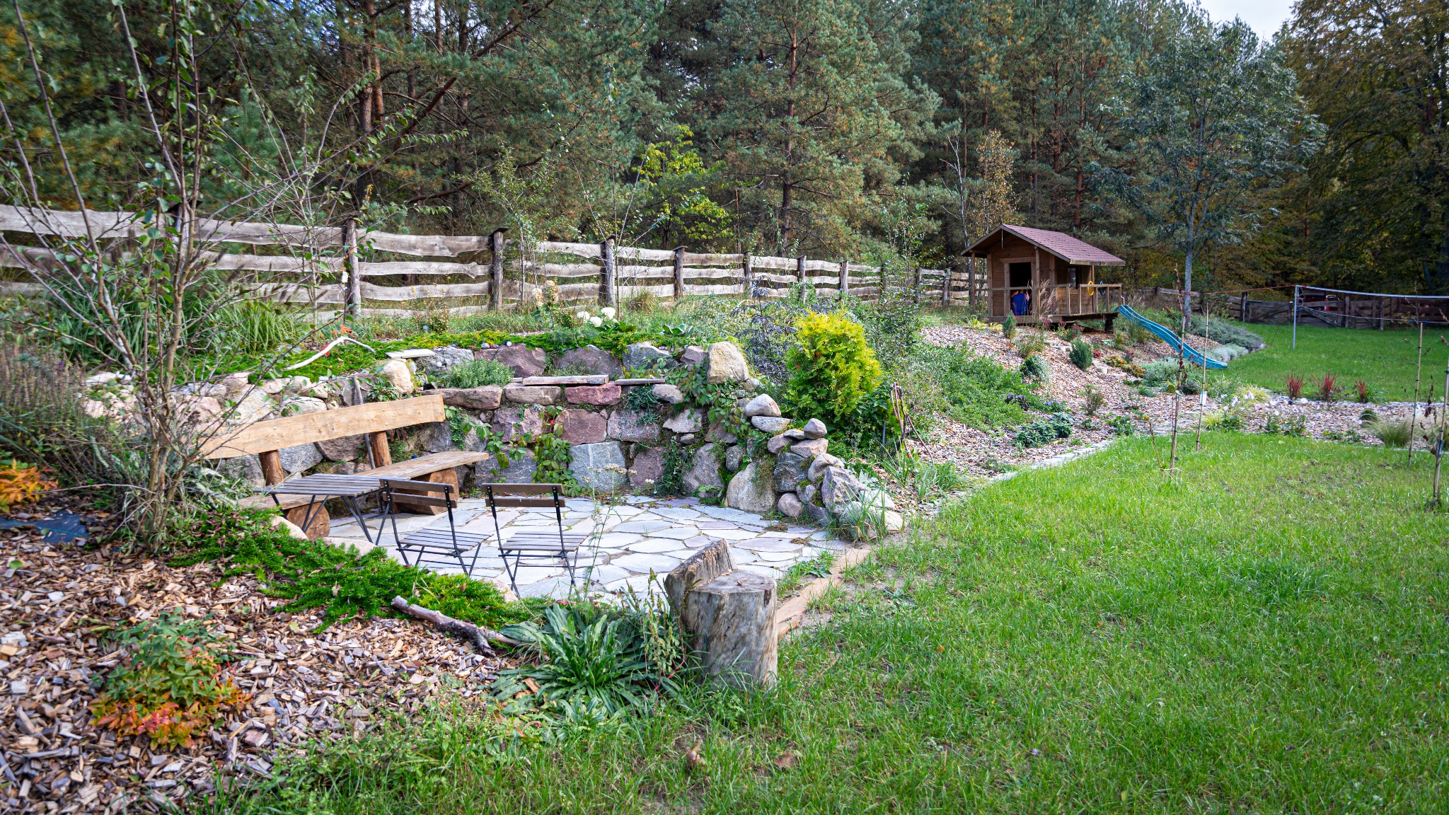 Ogród z drewnianym domkiem dla dzieci, kamiennym tarasem z meblami ogrodowymi, skalniakiem i drewnianym płotem w tle.