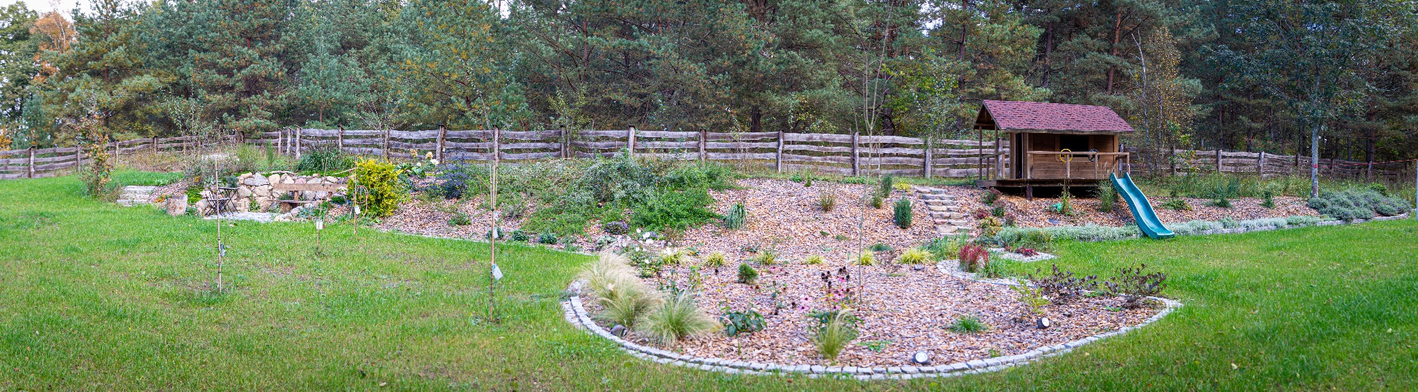 Szeroki kadr na zadbany ogród z drewnianą chatką dla dzieci ze zjeżdżalnią, rabatami wysypanymi korą i obrzeżami z kostki, drewnianym płotem w tle i drzewami.
