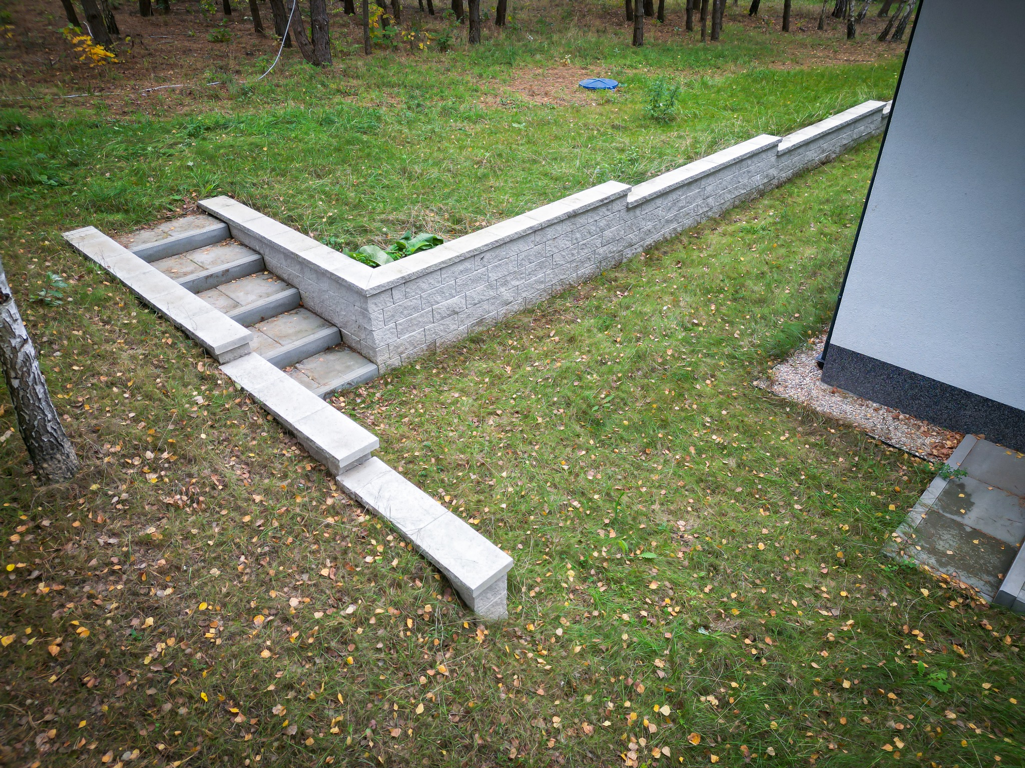 Nowoczesny murek oporowy z jasnoszarej kostki przy domu w Świętajnie, zintegrowany ze schodami prowadzącymi na trawnik pokryty opadłymi liśćmi.