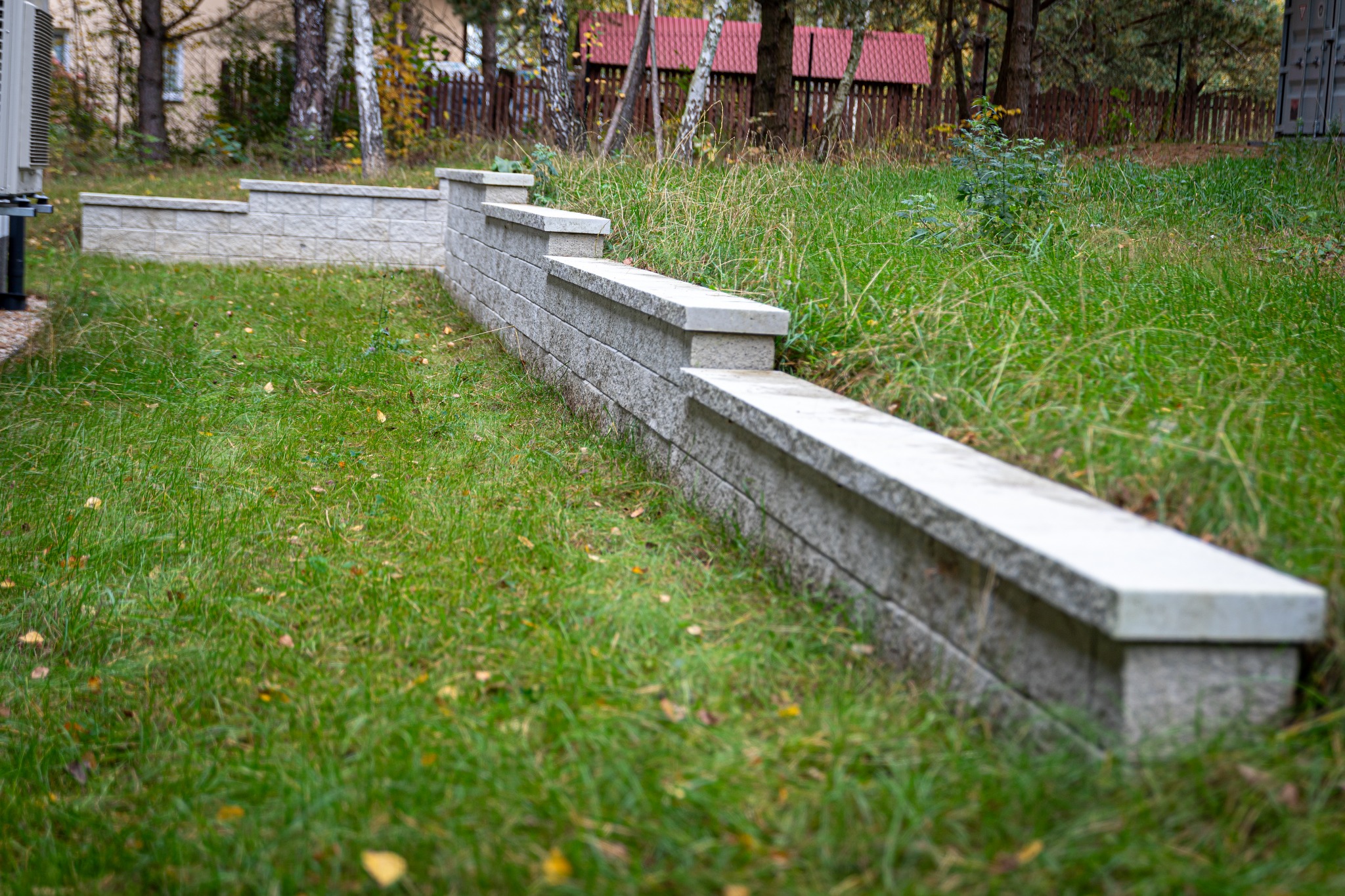Niska, modułowa ściana z szarej kostki betonowej tworząca obrzeże trawnika, widoczna z bliska w jesiennym ogrodzie z wysoką trawą i drzewami w tle.