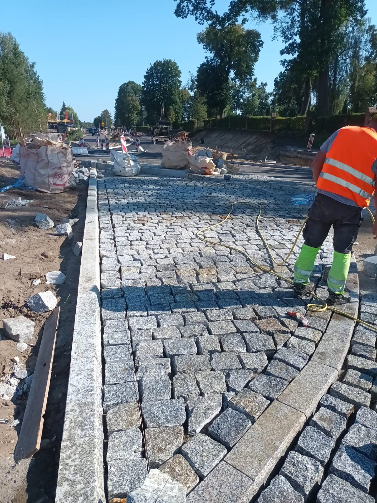 Układanie kostki granitowej na drodze. Widoczny fragment ułożonej nawierzchni, pracownik w kamizelce odblaskowej i worki z materiałem budowlanym w tle.