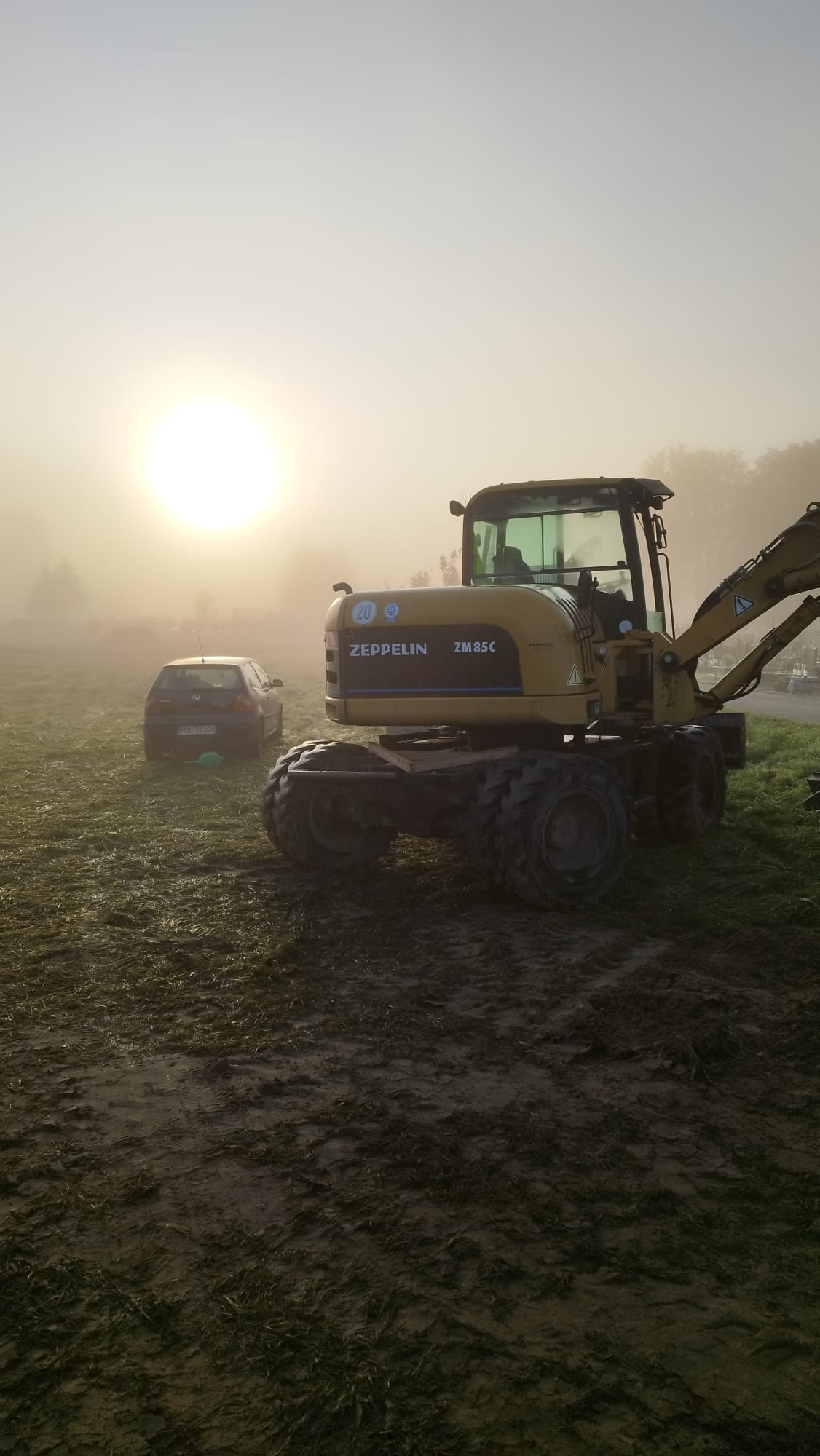 Żółta koparka Zeppelin ZM85C na polu w porannej mgle, obok zaparkowany samochód VW Golf. Słońce przebija się przez mgłę.