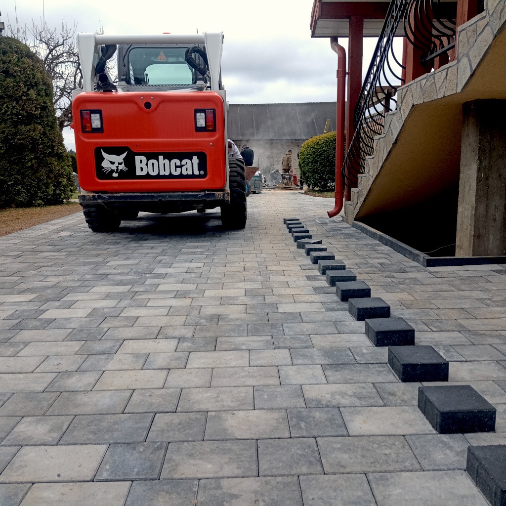 Ułożona kostka brukowa w odcieniach szarości z pomarańczowym Bobcatem w tle. Widoczne czarne kostki czekające na ułożenie wzdłuż ściany budynku.
