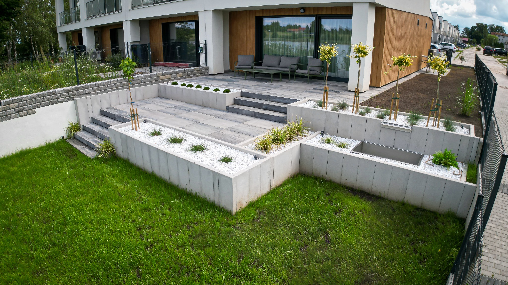 Nowoczesny ogród z tarasem wyłożonym szarą kostką, białymi kamieniami w rabatach i świeżo posadzoną trawą. Widok na budynek mieszkalny i ulicę.
