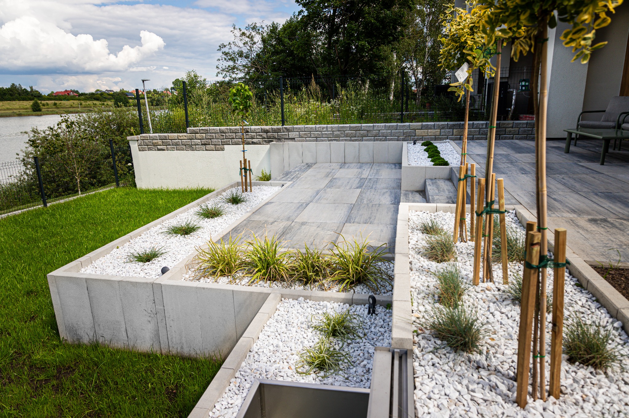 Nowoczesny ogród z szarymi płytami, białymi kamieniami i ozdobnymi trawami. Widok na jezioro w tle. Minimalistyczny design z elementami zieleni i kamienia.