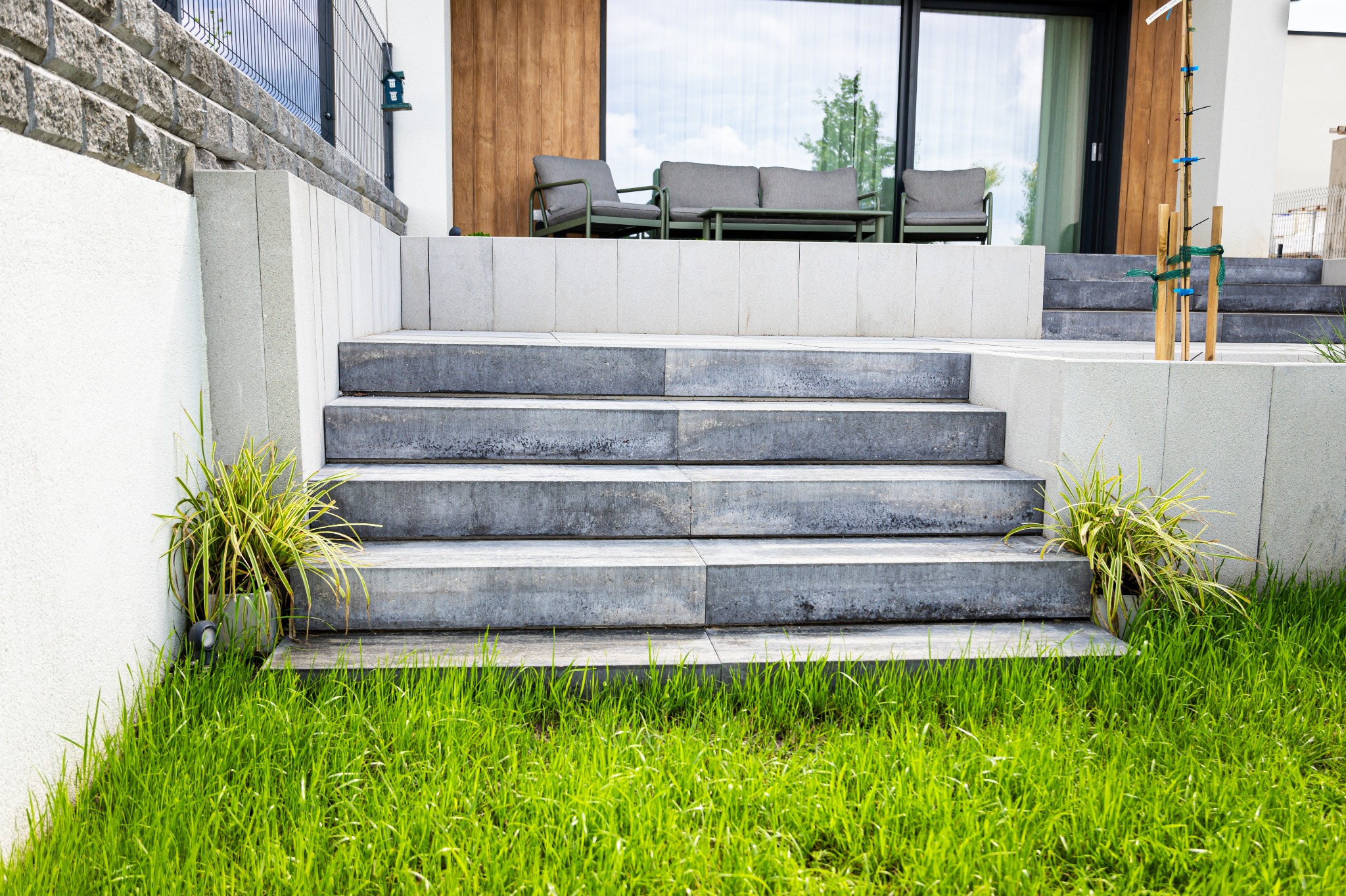 Schody z kostki brukowej prowadzące do nowoczesnego domu z zielonym trawnikiem i roślinami ozdobnymi. Widoczne meble ogrodowe na tarasie.