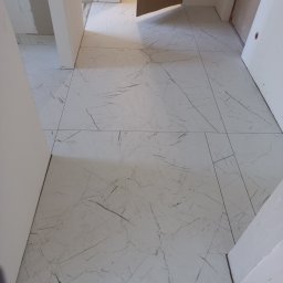 Robert Augusciak - Ułożona podłoga z płytek imitujących marmur w korytarzu, widoczne niedokończone prace wykończeniowe przy listwach przypodłogowych.