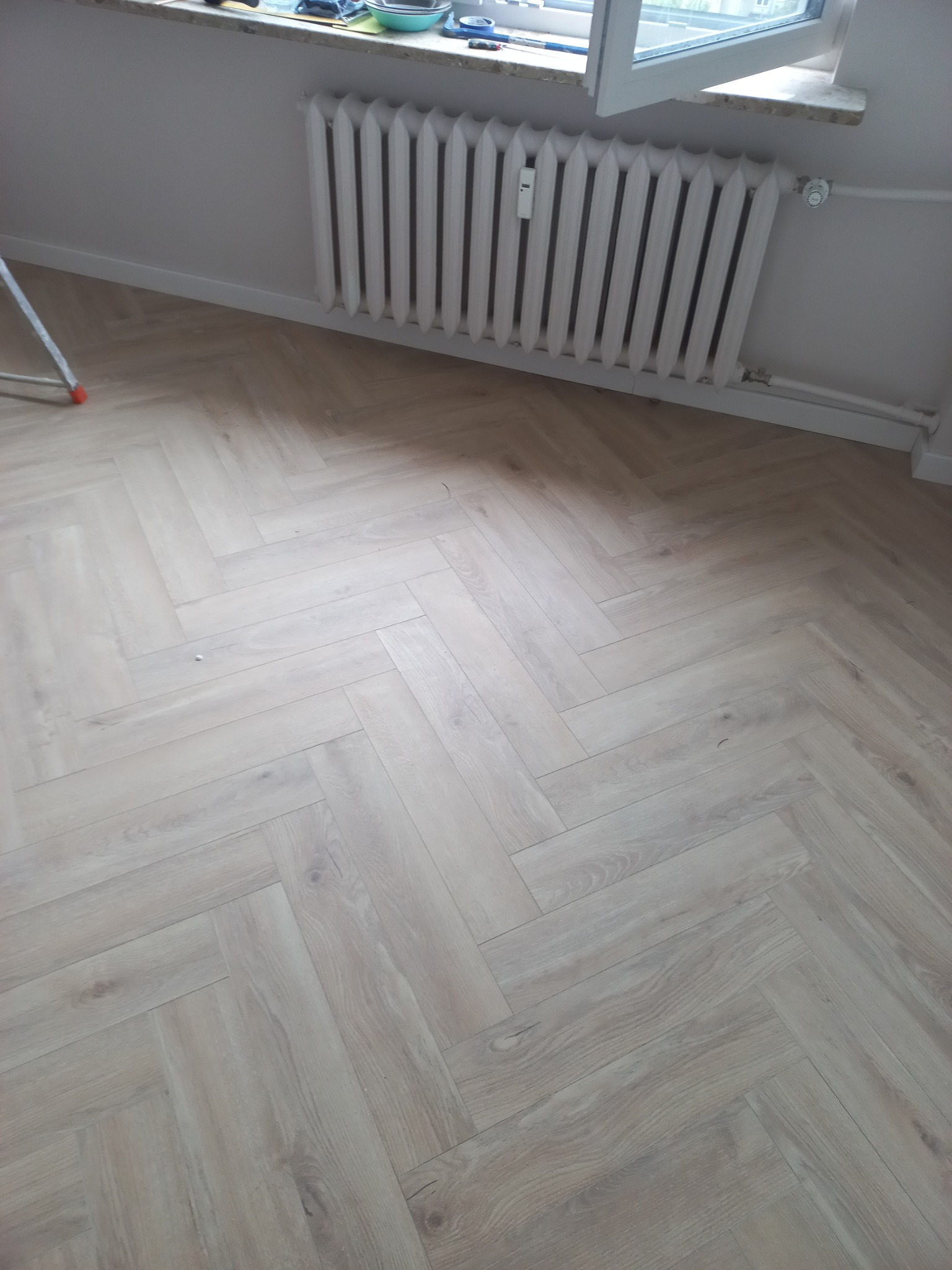 Jasna, nowa podłoga ułożona w jodełkę francuską, z widocznym białym grzejnikiem żeberkowym i fragmentem drabiny malarskiej w rogu pokoju.