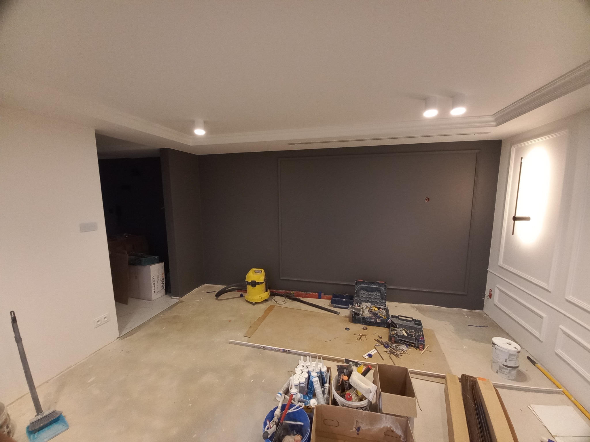 Wnętrze pokoju w trakcie wykańczania: szara ściana z dekoracyjną ramą, biała ściana z listwami, widoczne narzędzia, odkurzacz, puszki z klejem i farbą, przygotowane listwy przypodłogowe.