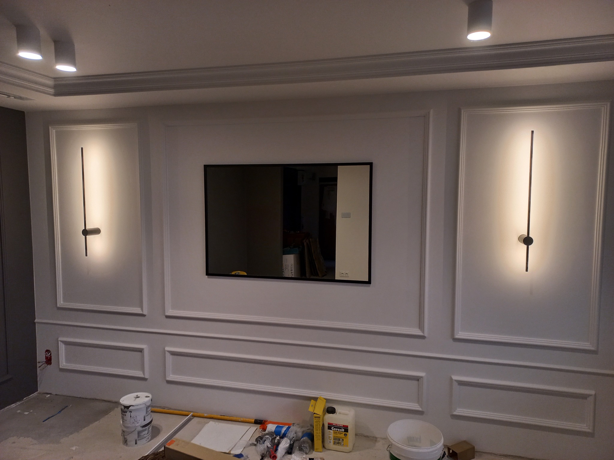 Ściana po malowaniu z dekoracyjnymi listwami, nowoczesnymi lampami ściennymi i wiszącym telewizorem, widoczne narzędzia malarskie i wiaderko z farbą na podłodze.