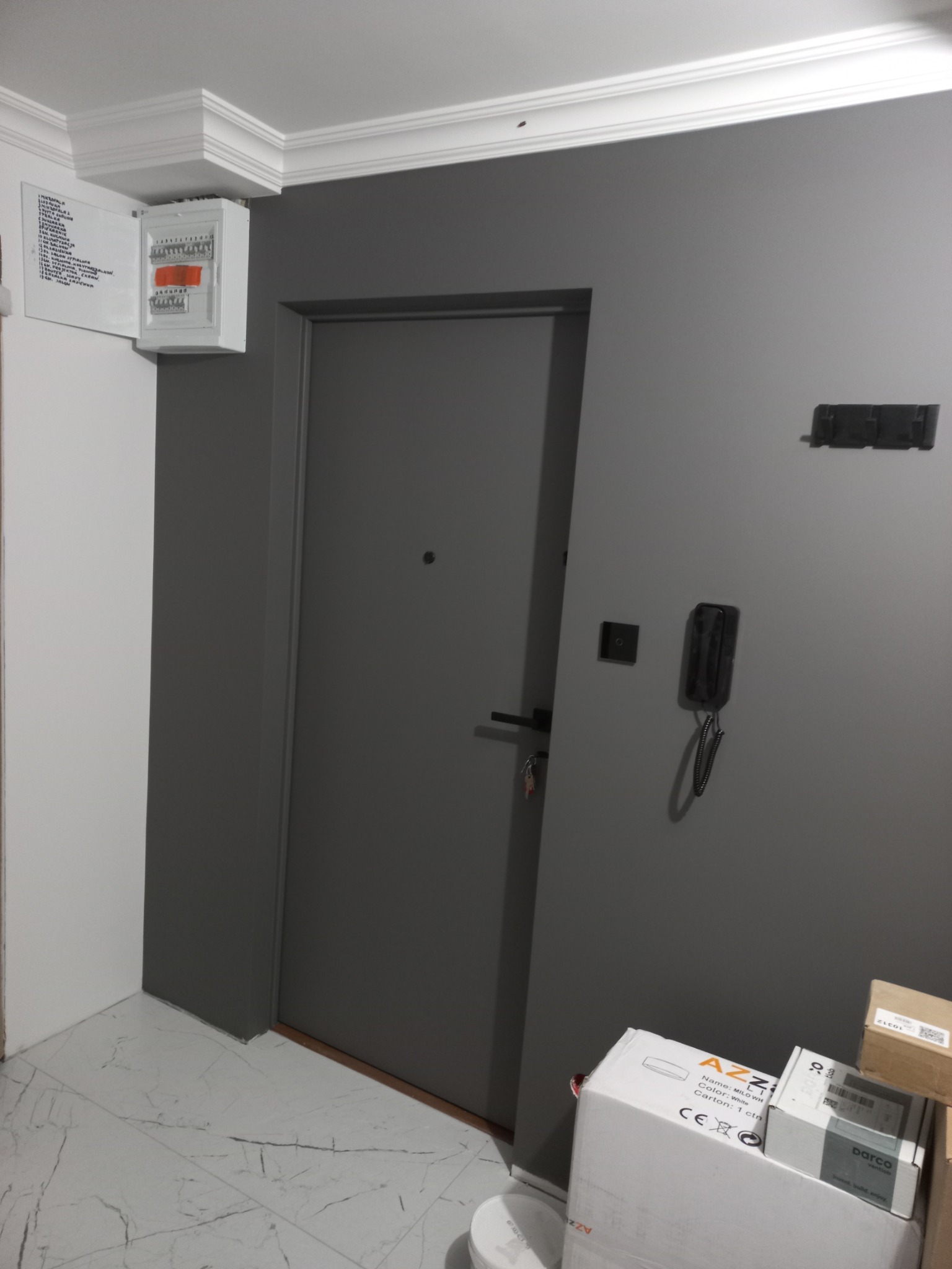 Szara ściana z zamontowaną skrzynką elektryczną z otwartymi drzwiczkami, obok drzwi wejściowe, domofon i wieszak na ubrania; na podłodze kartony z akcesoriami.
